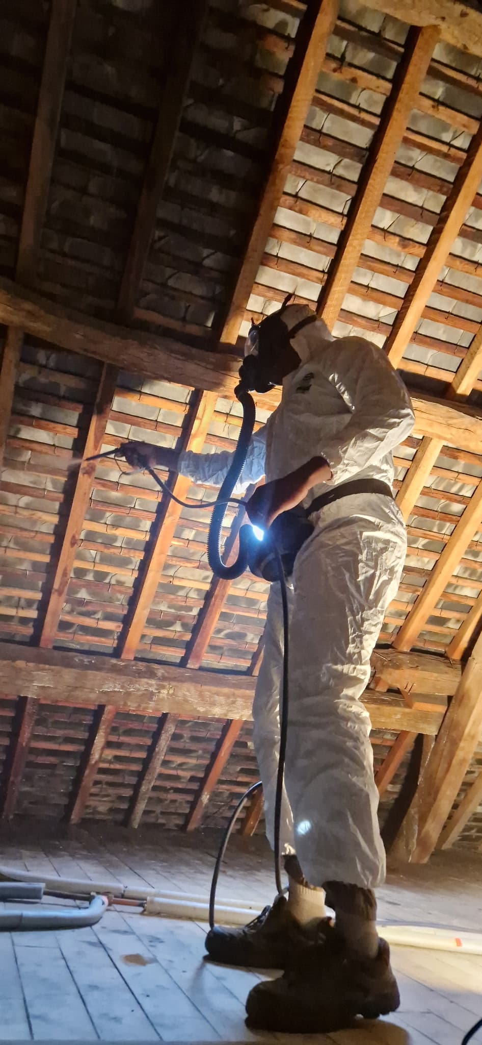 Technicien ACO-HABITAT pulvérisant un produit de traitement sur une charpente, en combinaison et masque de protection